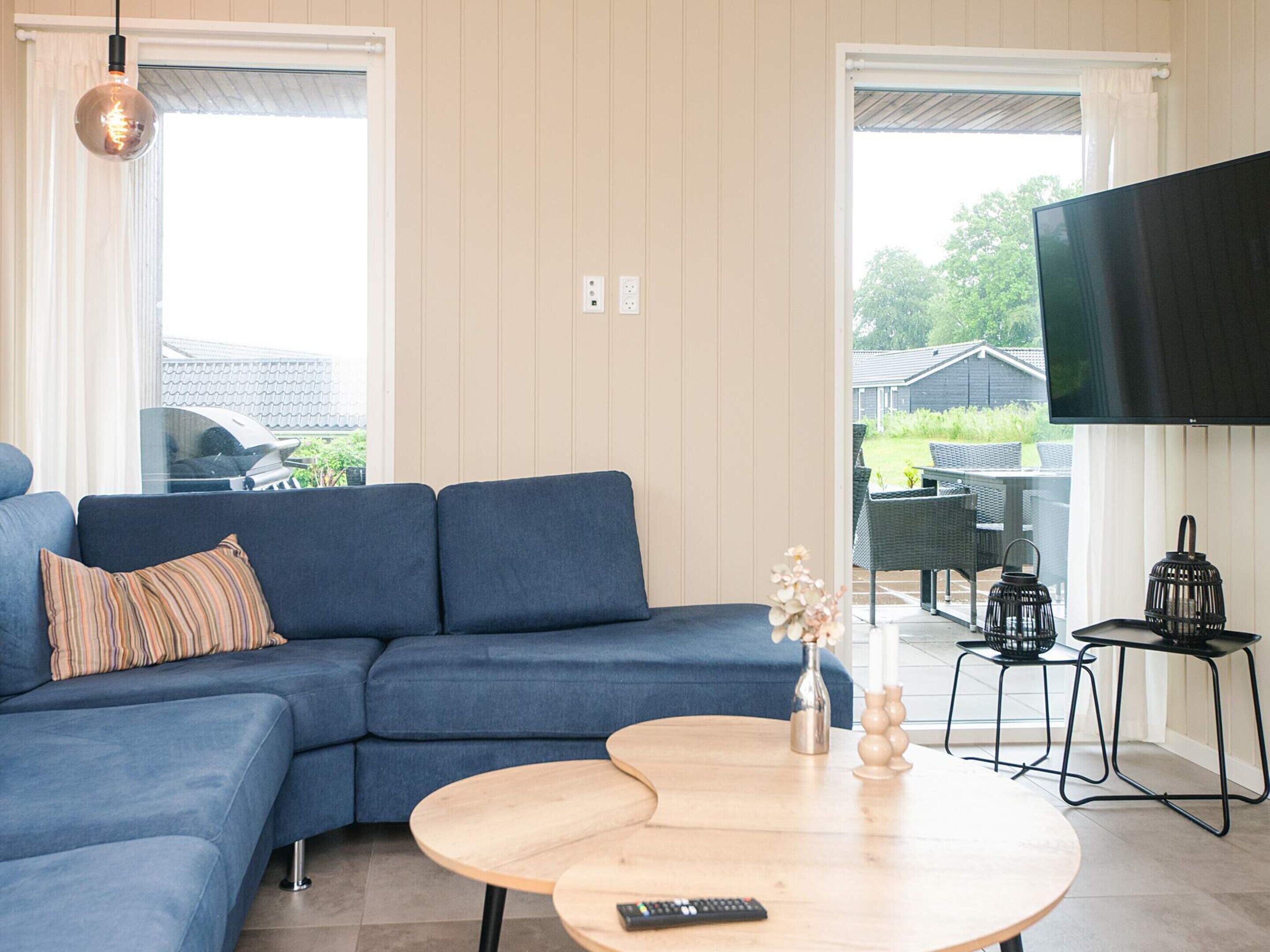 A living room with a blue sofa, a round wooden coffee table, a TV mounted on the wall, and a view of the outside.