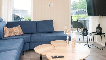 A living room with a blue sofa, a round wooden coffee table, a TV mounted on the wall, and a view of the outside.