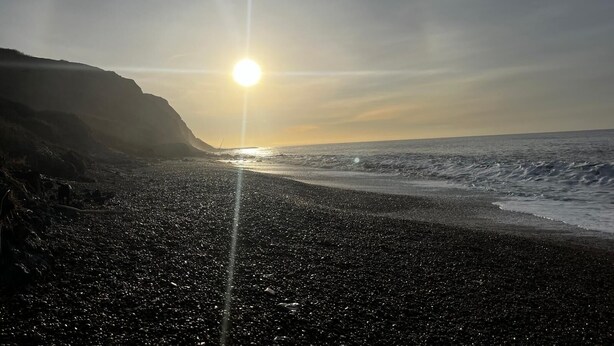 Une plage où le soleil se couche sur l’océan, avec des falaises rocheuses et un ciel dégagé.