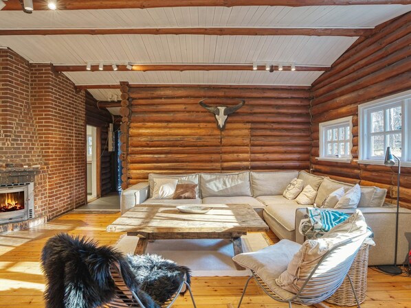 Interior de una cabaña de troncos con chimenea de ladrillo, un conjunto de sofás, una mesa de comedor y una silla de mimbre.