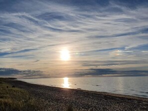 A beach with the sun setting over the water, a rocky shoreline, and a clear sky.