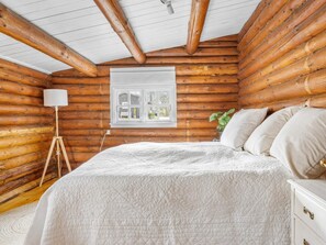 Ein Schlafzimmer in einer Blockhütte mit Bett, Nachttisch, Lampe, Fenster und Blick ins Freie.