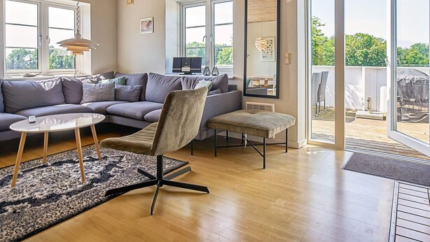 A modern living room with a grey sofa, a small round table, and a chair. There is a large mirror on the wall and a sliding glass door leading to an outdoor area. - 12 Person Holiday Home in Aakirkeby (Akirkeby)