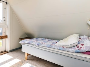 A small, single-bed room with a sloped ceiling, a wooden floor, and a window with a view of the outside.