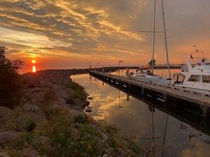 A marina at sunset with boats docked, a pier extending into calm waters, and a rocky shoreline with sparse vegetation.