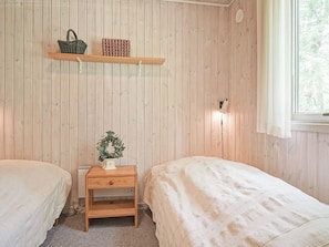 A room with two beds, a nightstand with a lamp, a shelf with books and a basket, and a window with curtains.