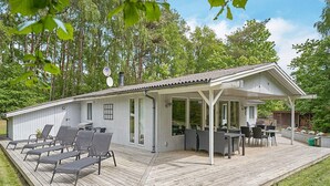 Une terrasse extérieure couverte, dotée d’un espace salon et d’un coin repas, le tout entouré d’arbres.