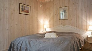 Un dormitorio con pared revestida de madera, una cama con cubrecama gris, dos lámparas de noche y dos cuadros enmarcados.