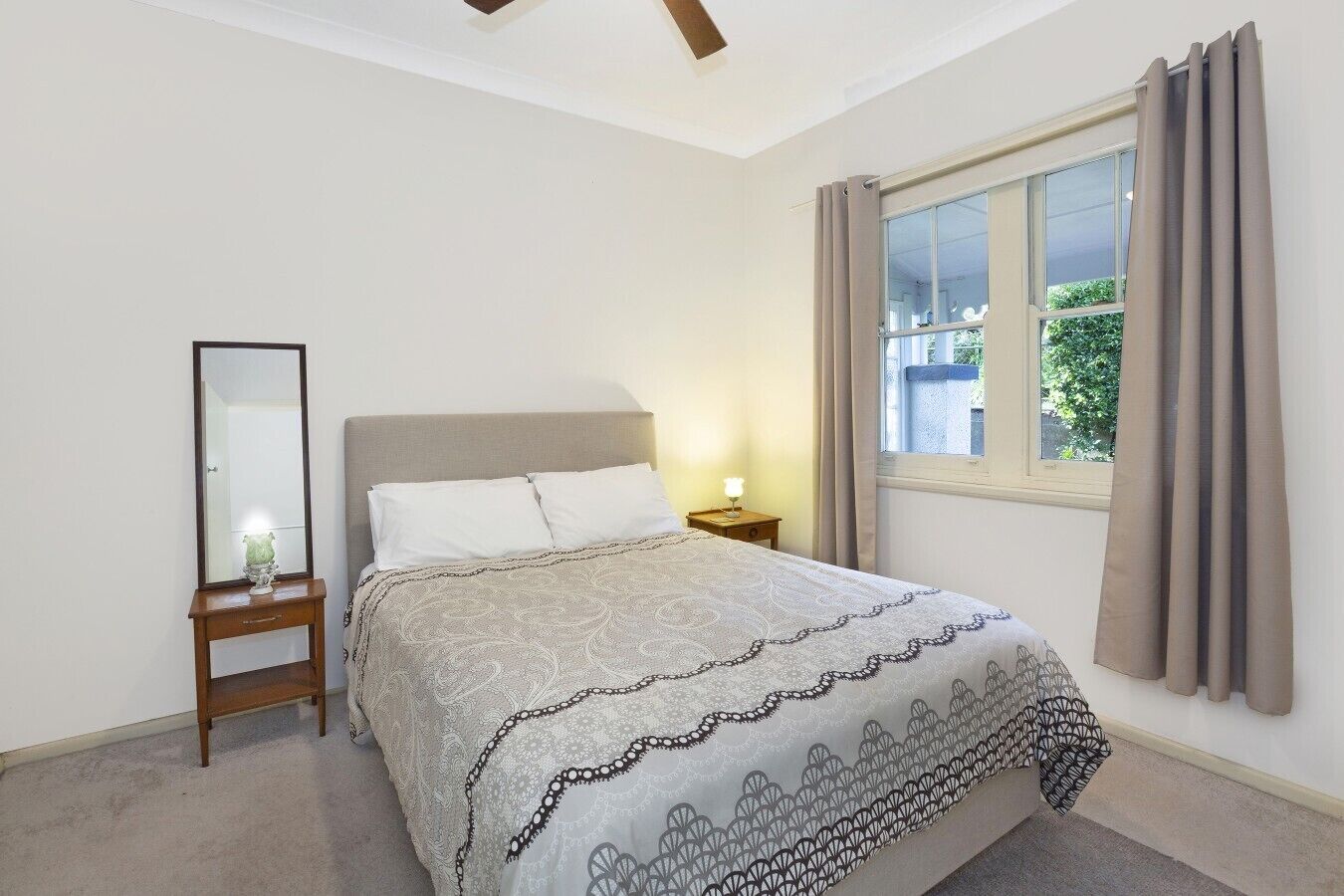 A bedroom with a bed, a nightstand, a mirror, and a window with curtains.