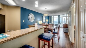 A modern kitchen with a granite countertop, wooden bar stools, and a large clock on the wall. - 🌞Oceanfront | Indoor Heated Water Park + Lazy River | Dunes Village Resort 1120 (Myrtle Beach)