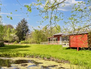 A red cabin with a deck, surrounded by a grassy field and a pond. - 4 Person Holiday Home in Hojslev-by Traum (Hojslev)