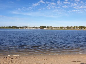 A calm lake with boats, a sandy beach, and a distant shoreline with buildings and trees.