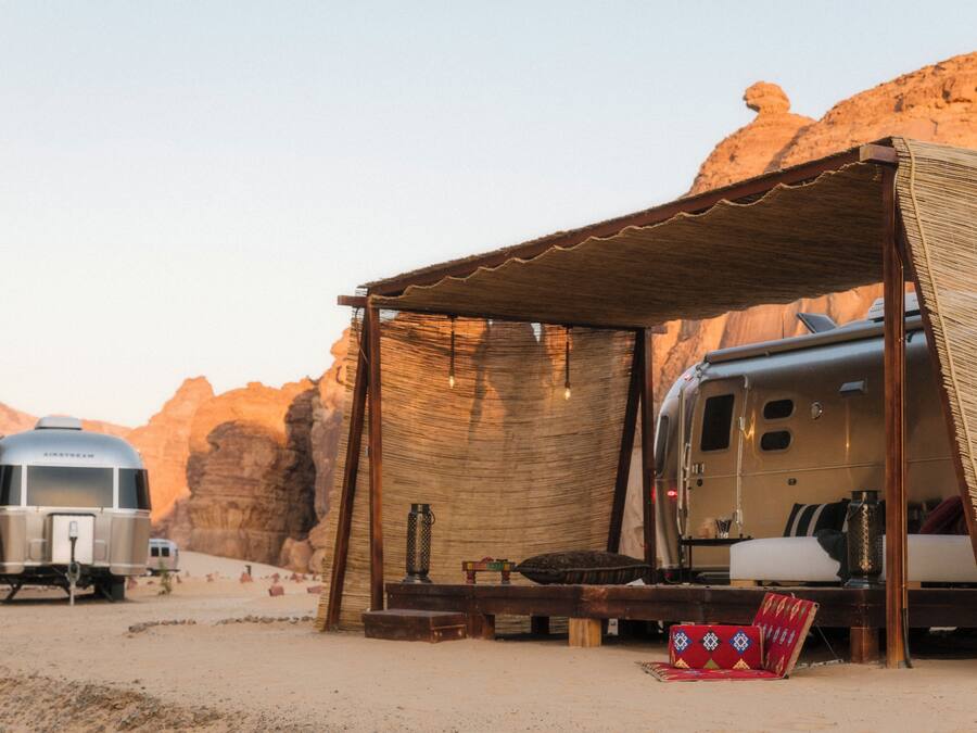 Una caravana con un espacio exterior cubierto, una cama y una pequeña mesa con una lámpara.