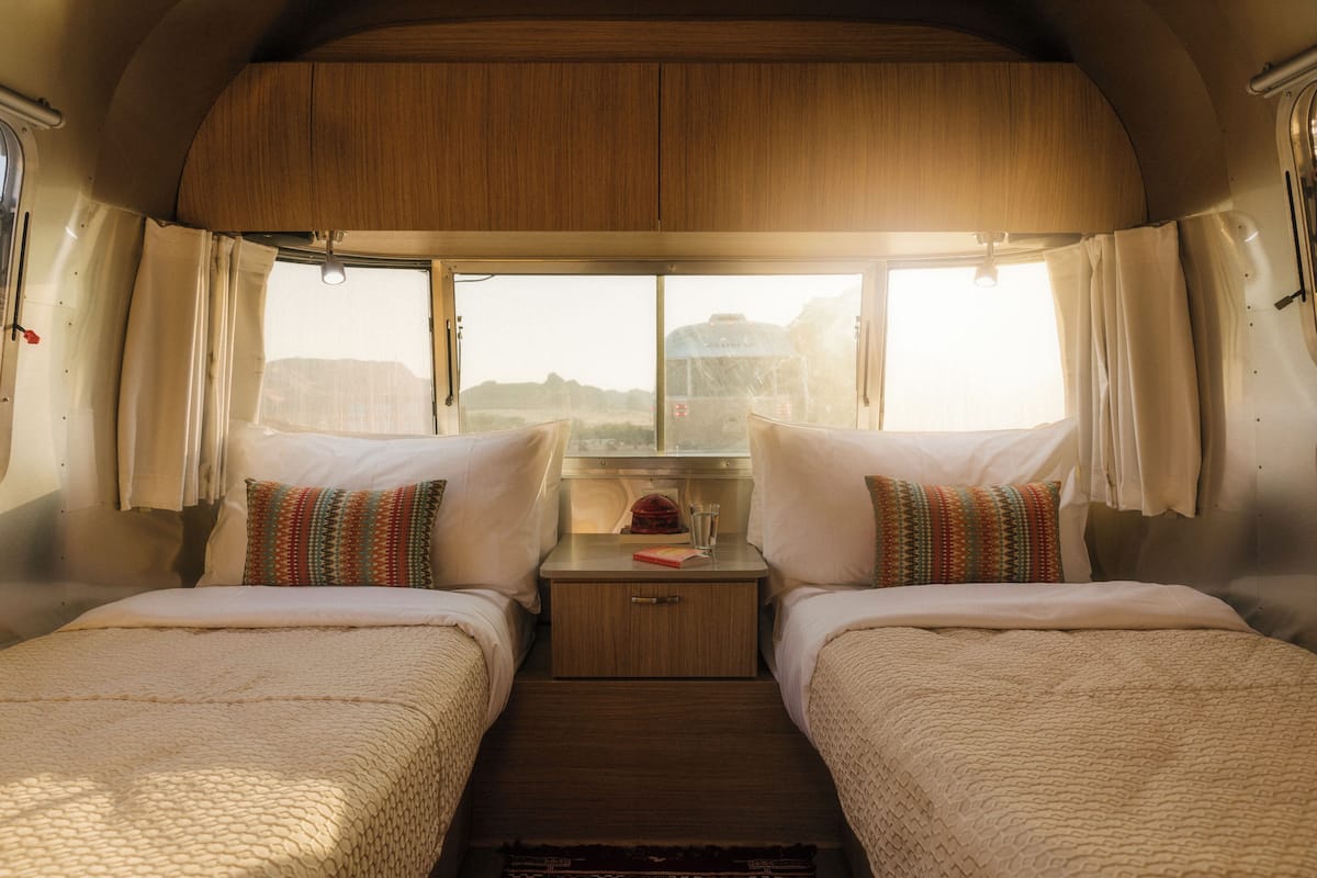 A cozy RV bedroom with two beds, a small table with a book and glasses, and a window with curtains.