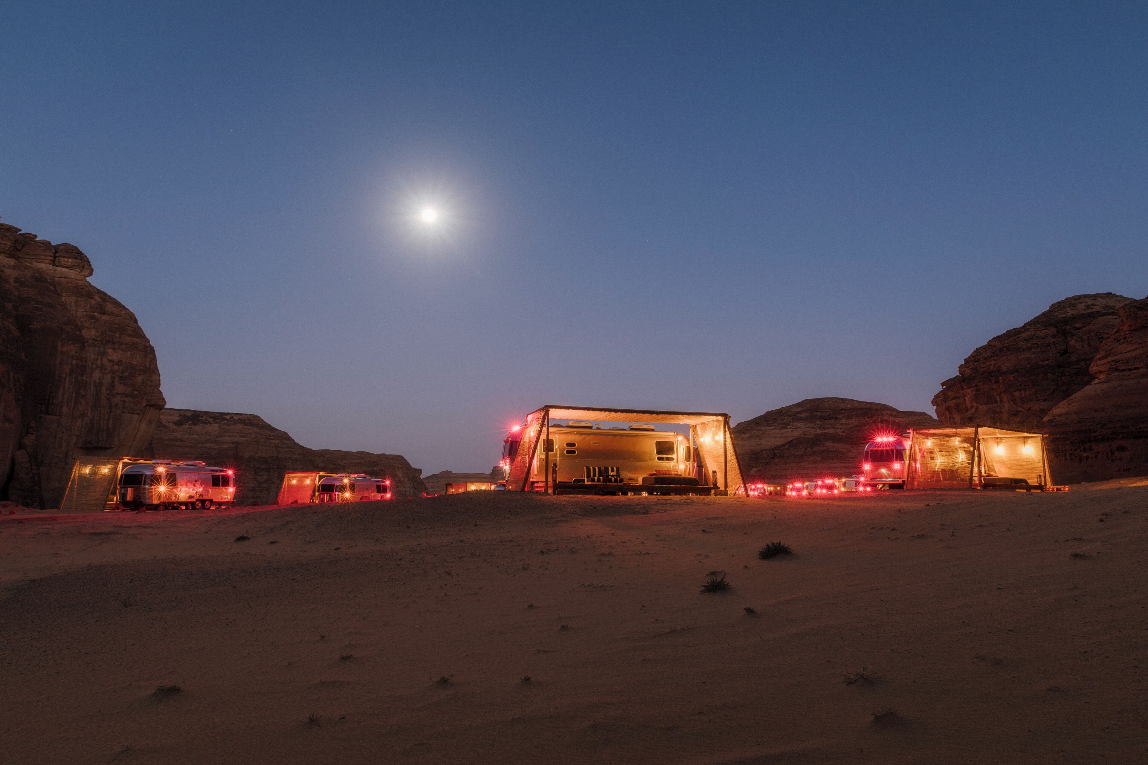 A desert campsite at dusk with a large, illuminated tent in the center, surrounded by smaller tents and vehicles.
