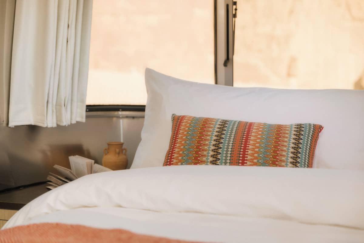A bed with a white pillow and a colorful patterned pillow, a vase, and a window with curtains.