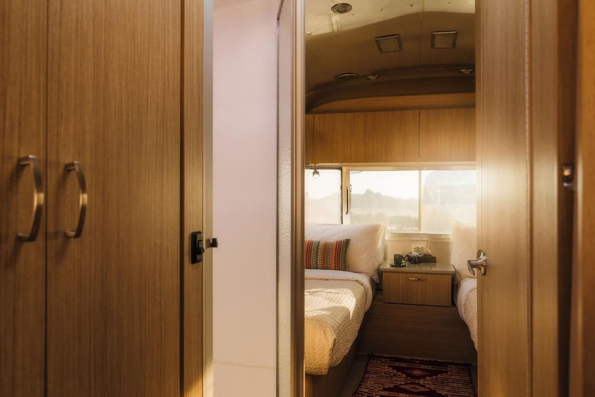 A compact, well-lit room with wooden cabinetry, a bed with a colorful pillow, a small bedside table, and a window providing a view of the outdoors.