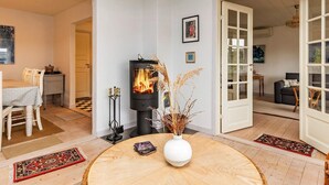 A cozy living room with a wood-burning stove, a dining area with a table and chairs, and a view into another room through an open door. - 5 Person Holiday Home in Laeso (Læsø)