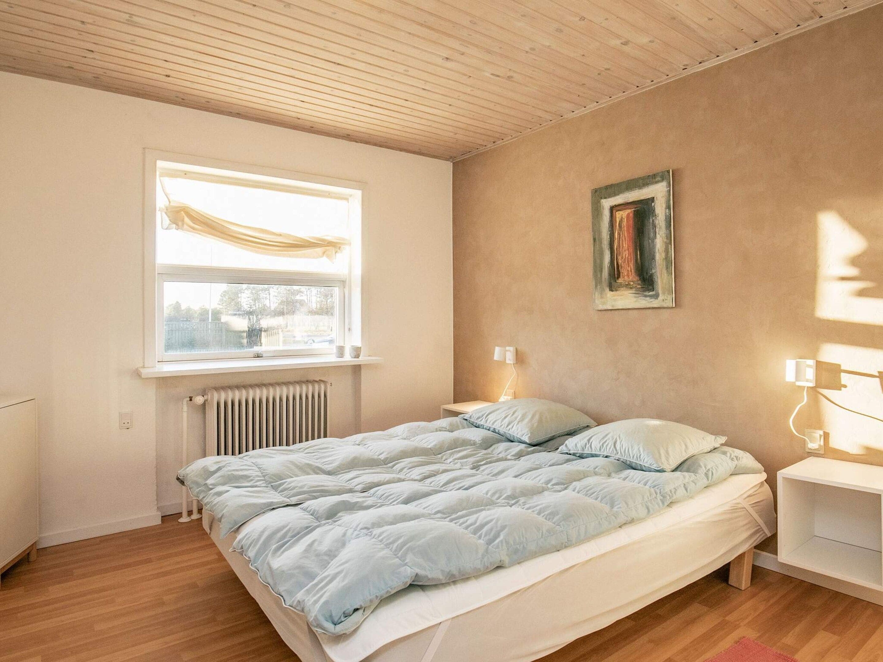 Et soveværelse med seng, et vindue med gardiner, en radiator, et sengebord med lamper og en væg med et billede i ramme.