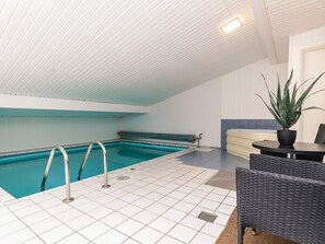 Una piscina interior con suelo de baldosas, una escalera metálica y una zona de estar con mesa y sillas.