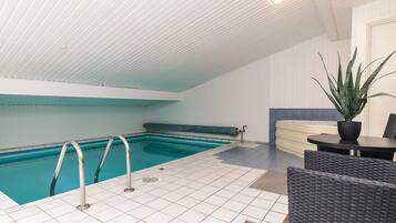 En indendørs swimmingpool med flisegulv, en metalstige og et opholdsområde med et bord og stole.