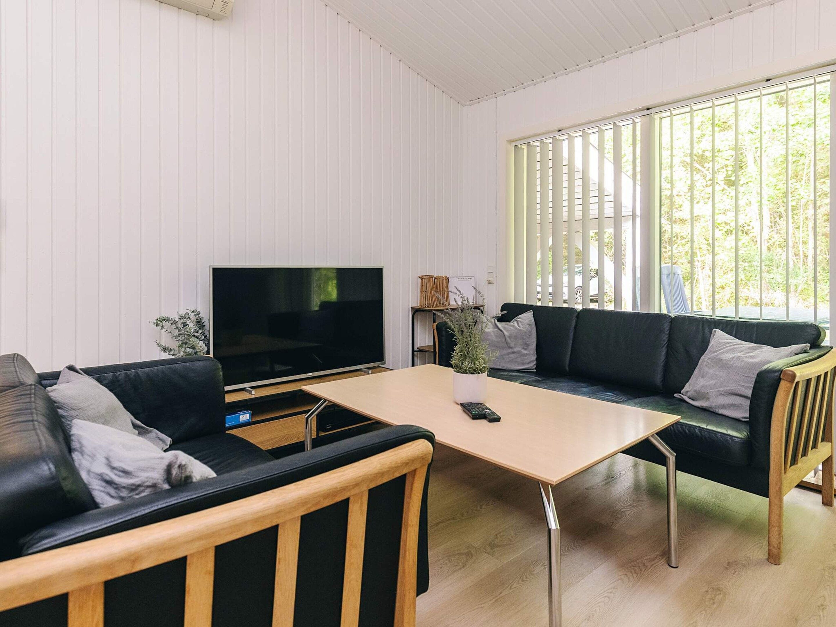 En moderne stue med et fladskærms-tv, to sorte lædersofaer, et træ sofabord og et stort vindue med lodrette skodder.