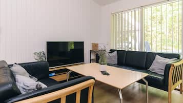 En moderne stue med et fladskærms-tv, to sorte lædersofaer, et træ sofabord og et stort vindue med lodrette skodder.