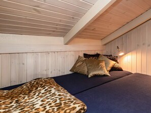 A room with a sloped ceiling, wooden panel walls, a bed with a dark blue mattress and leopard print blanket, and a single bedside lamp.