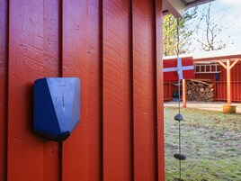 Una estructura de madera roja con un objeto azul montado, un cartel rojo y blanco, y una pila de leña al fondo.