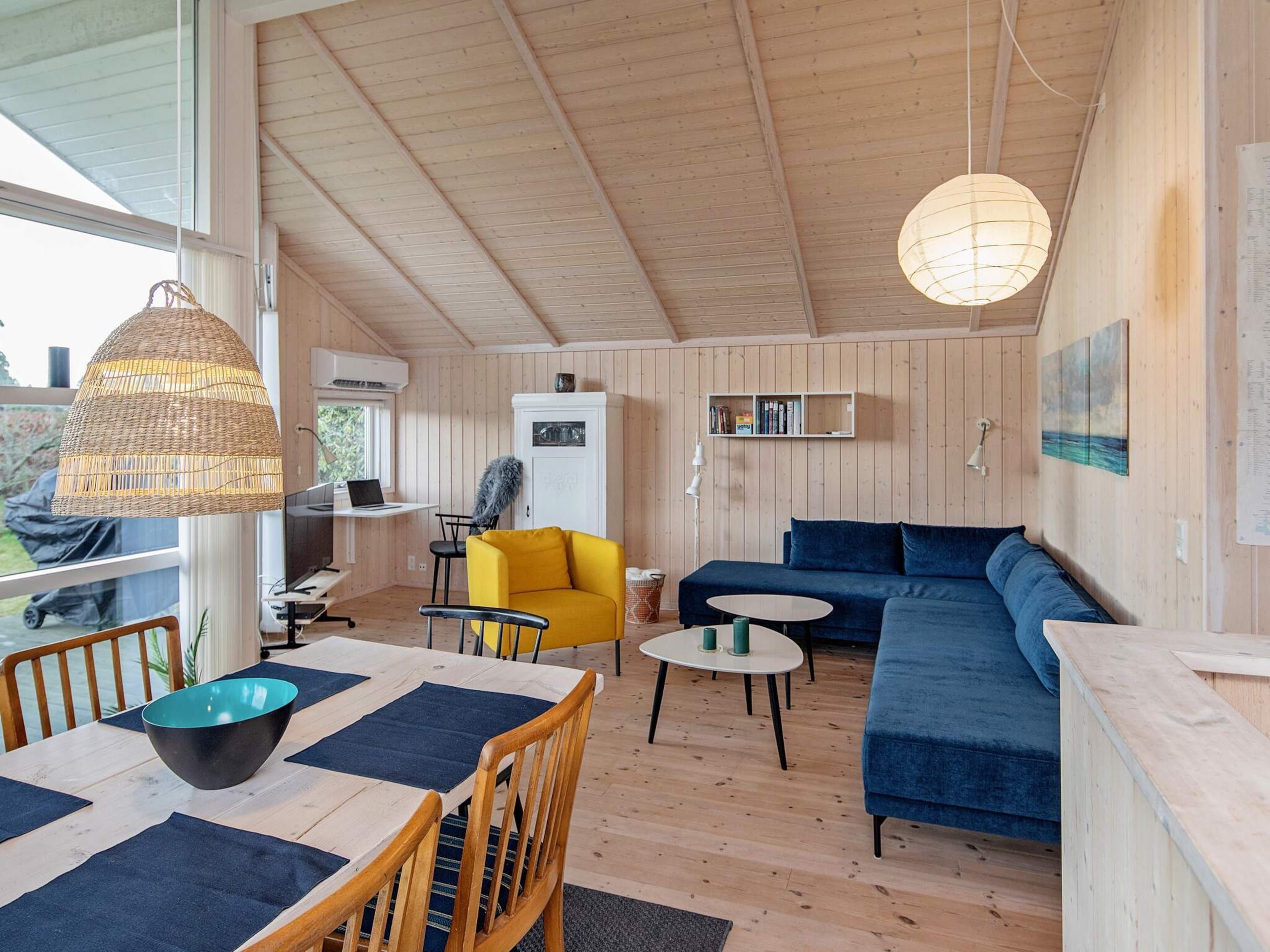 A wooden interior with a dining table, yellow armchair, blue sofa, and pendant lighting.