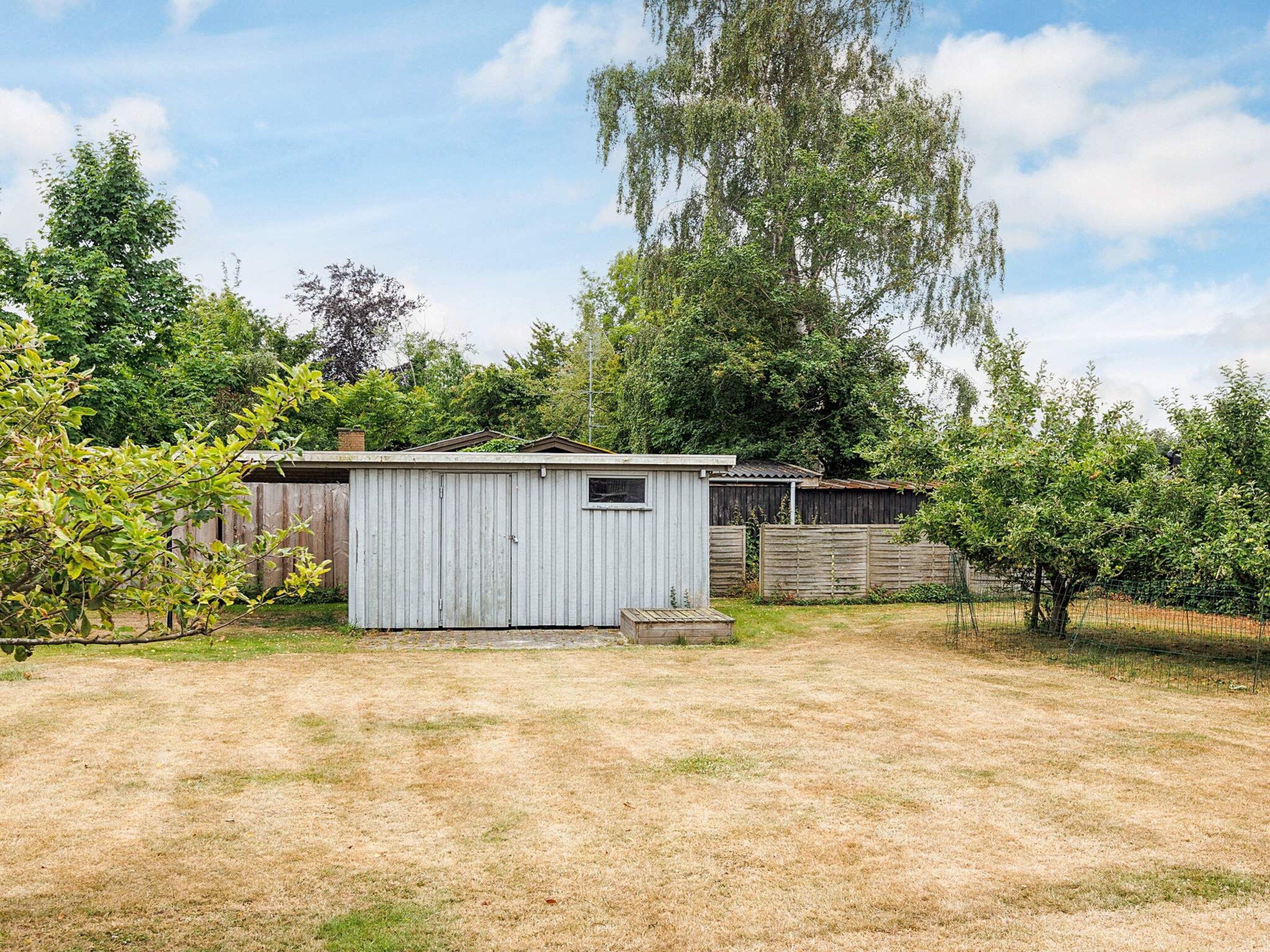 A small, single-story shed with a sloped roof, surrounded by a wooden fence and trees.