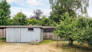 A small, single-story shed with a window and a door, surrounded by a fenced yard with trees and a wooden fence in the background. - 4 Star Holiday Home in Vaeggerlose (Vaeggerlose)