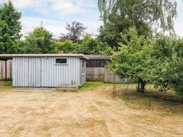A small, single-story shed with a window and a door, surrounded by a fenced yard with trees and a wooden fence in the background.