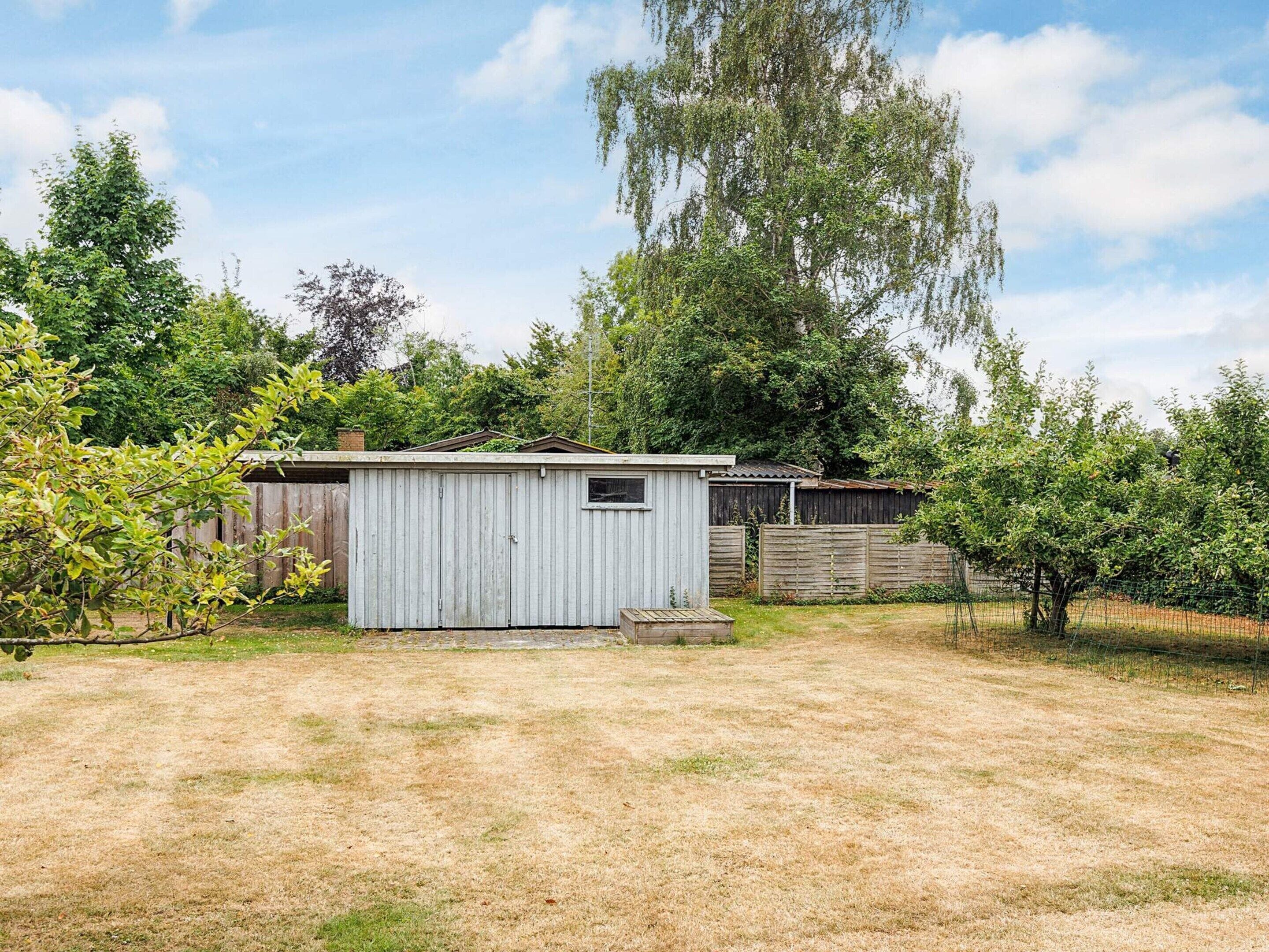 A small, single-story shed with a sloped roof, surrounded by a fenced yard with sparse grass and trees.