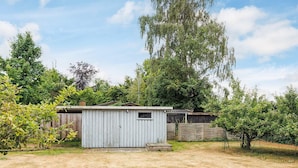 A small, single-story shed with a sloped roof, surrounded by a fenced yard with sparse grass and trees. - 4 Star Holiday Home in Vaeggerlose (Vaeggerlose)