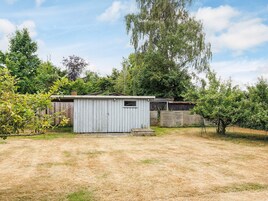 A small, single-story shed with a sloped roof, surrounded by a fenced yard with sparse grass and trees.