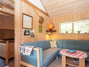 A cozy cabin interior with a blue sofa, a small wooden table with a checkered tablecloth, and a window with a view of greenery.