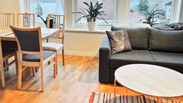 Una sala de estar moderna con comedor, ventanas amplias que dan a un lago y un sofá gris con un cojín decorativo.
