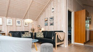 Un salón acogedor con un sofá de cuero negro, una mesa de centro redonda y un reposapiés gris. La habitación tiene paredes de madera y un techo alto con vigas a la vista.