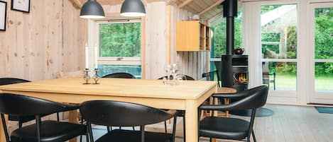 A wooden interior with a dining table, chairs, pendant lights, a fireplace, and large windows.
