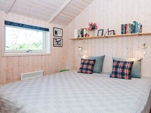 A room with a bed, two pillows, a window, a radiator, and a shelf with books and flowers. - 6 Person Holiday Home in Hemmet (Hemmet)