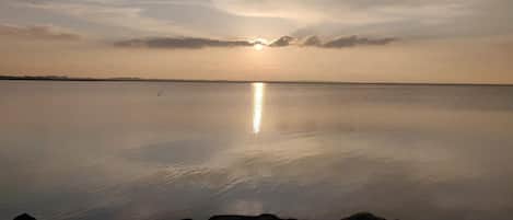 A calm body of water with a rocky shoreline in the foreground and a sunset in the background.