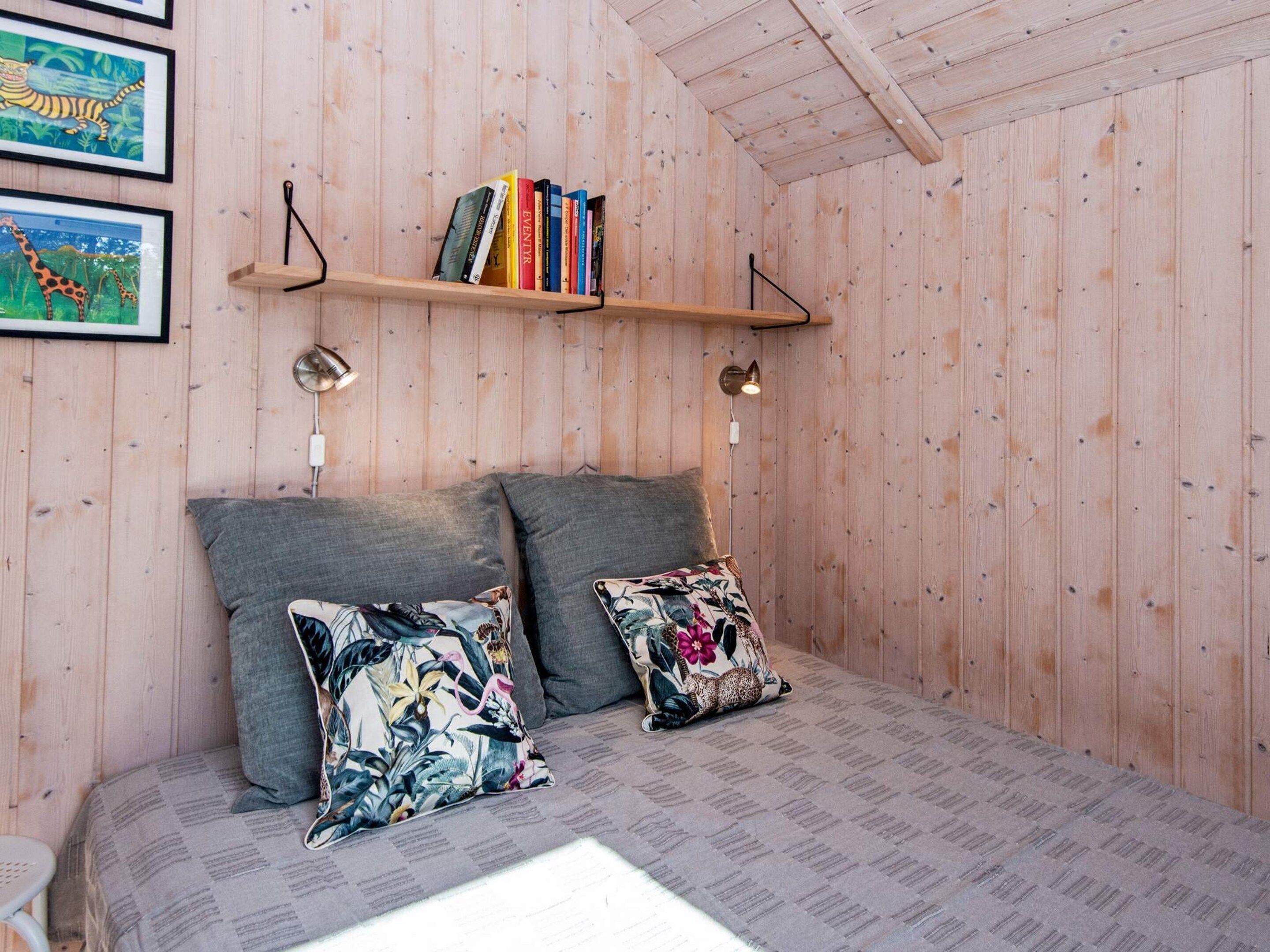 Ein gemütliches Zimmer mit einem Bett, zwei Kissen, einem Regal mit Büchern und gerahmten Bildern an der Wand.