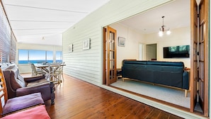 A spacious living area with a wooden floor, a dark green sofa, and a dining table with chairs. A television is mounted on the wall, and there is a view of the ocean through large windows. - 12 Bluewater Drive Narooma (Narooma)