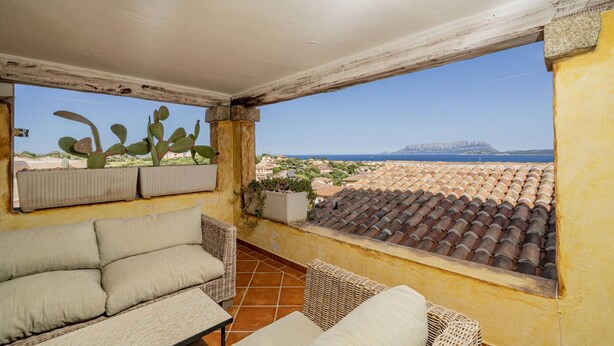 A balcony with wicker furniture, a cactus plant, and a view of a coastal town. - Casa Nadia by Wonderful Italy (Olbia)