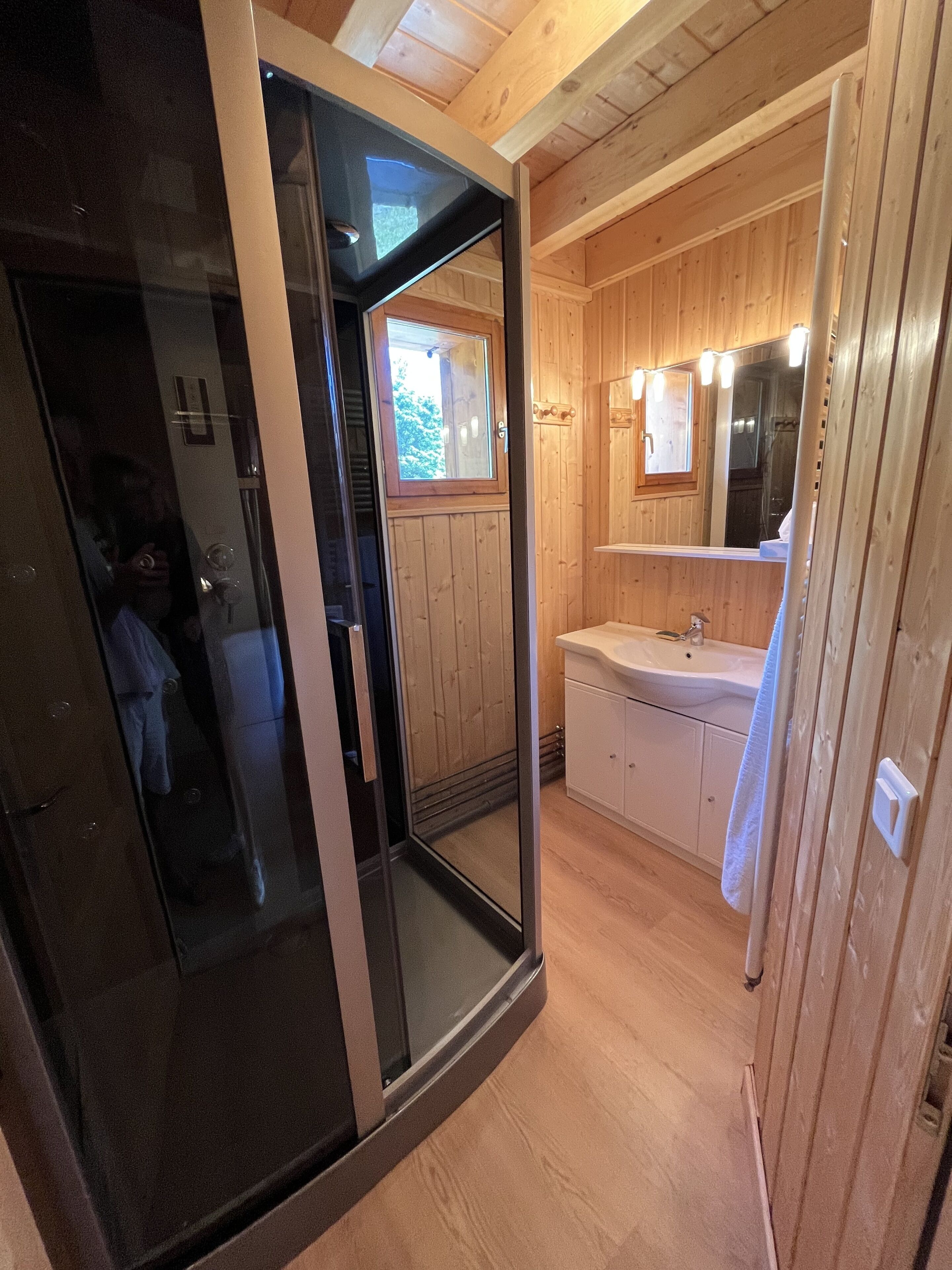 A wooden cabin interior with a glass door leading to a balcony, a bathroom with a sink and mirror, and wooden walls.