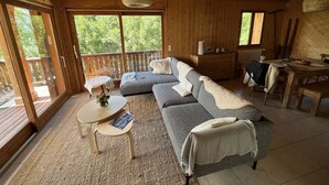 A cozy cabin interior with a wooden ceiling, a sofa, a dining table, and a large window offering a view of greenery. - Chalet + sauna with panoramic views in beautiful surroundings (Albiez-le-Jeune)