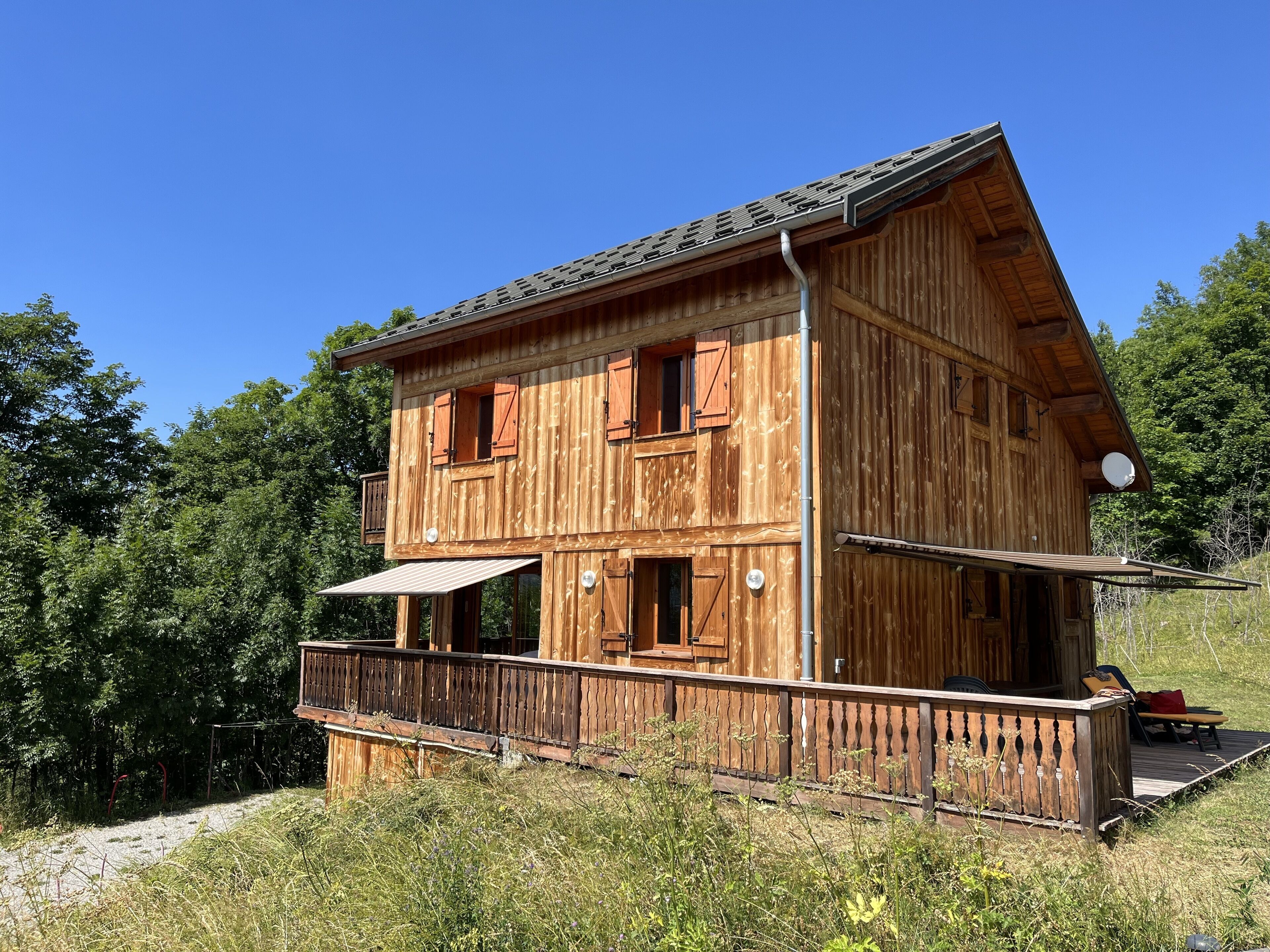 A wooden two-story house with a balcony, surrounded by greenery.