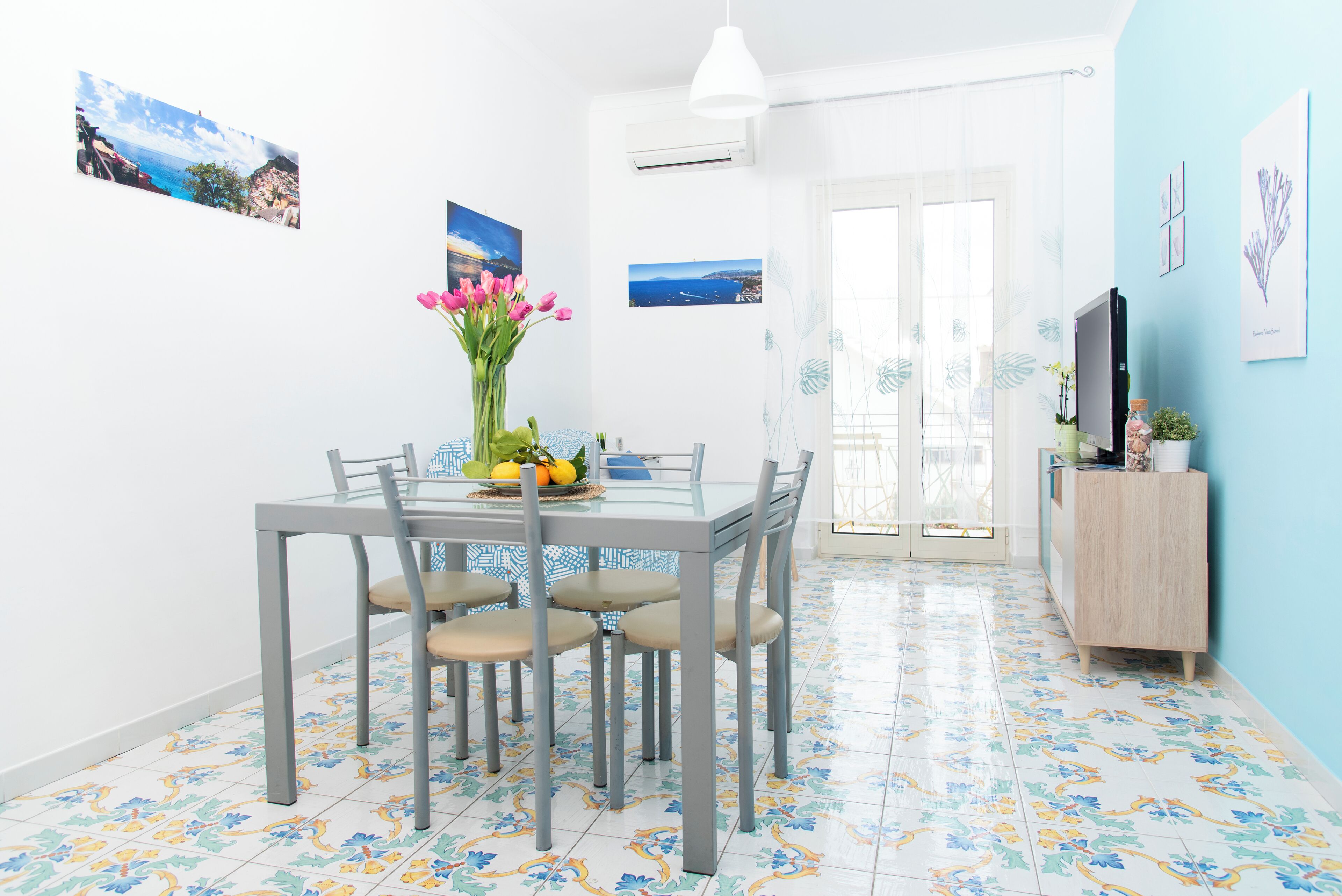 A dining area with a glass table, four chairs, a vase with flowers, and a fruit bowl. There are wall-mounted pictures and a TV on a wooden cabinet.