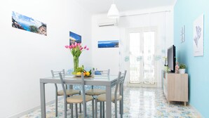 A dining area with a glass table, four chairs, a vase with flowers, and a fruit bowl. There are wall-mounted pictures and a TV on a wooden cabinet. - Luigi's room Sorrento (Meta)